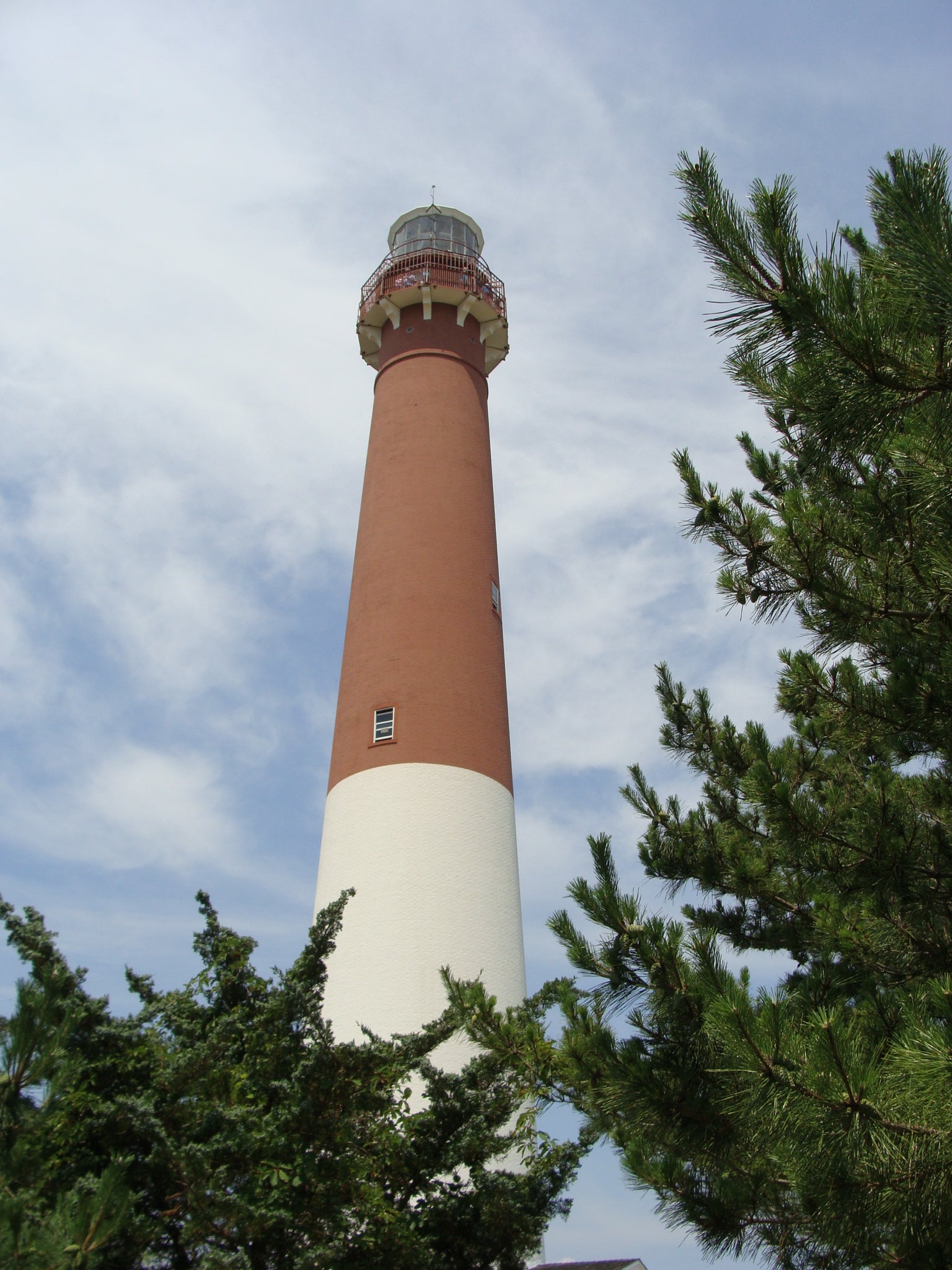 Lighthouse with a white top and brown body, surrounded by trees against a blue sky.