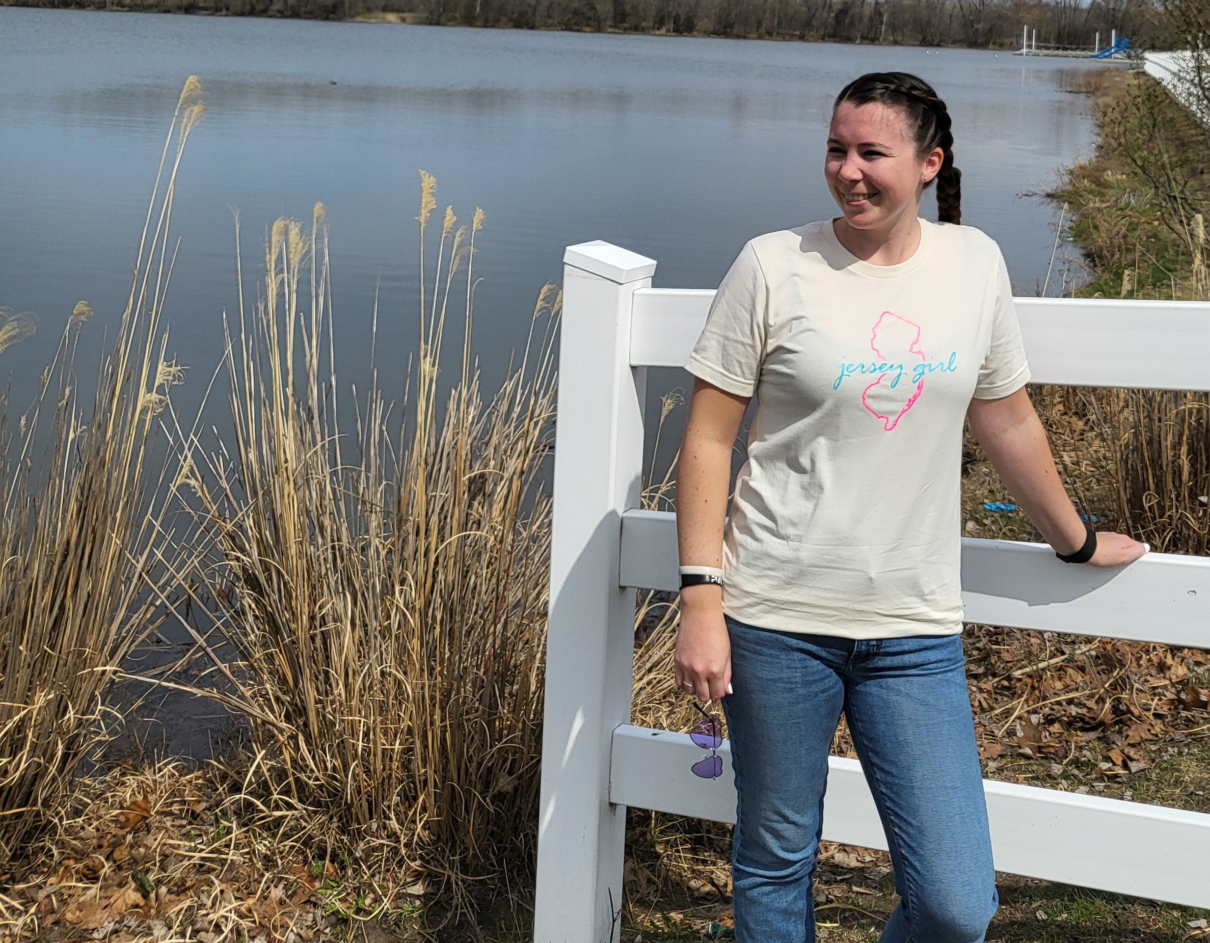 Woman standing by a white fence near a body of water with tall grasses.