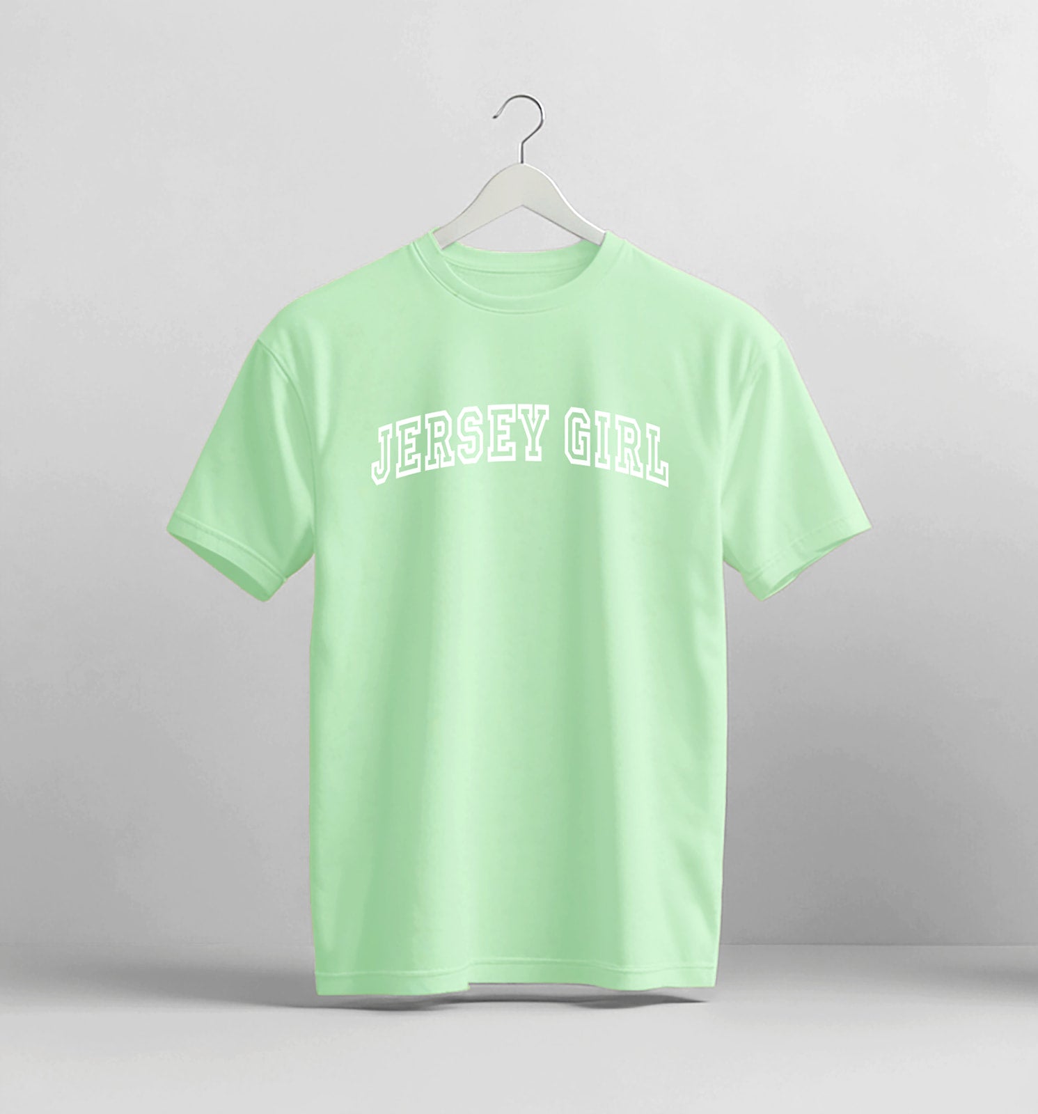 Light green t-shirt with 'Jersey Girl' text on a hanger against a gray background