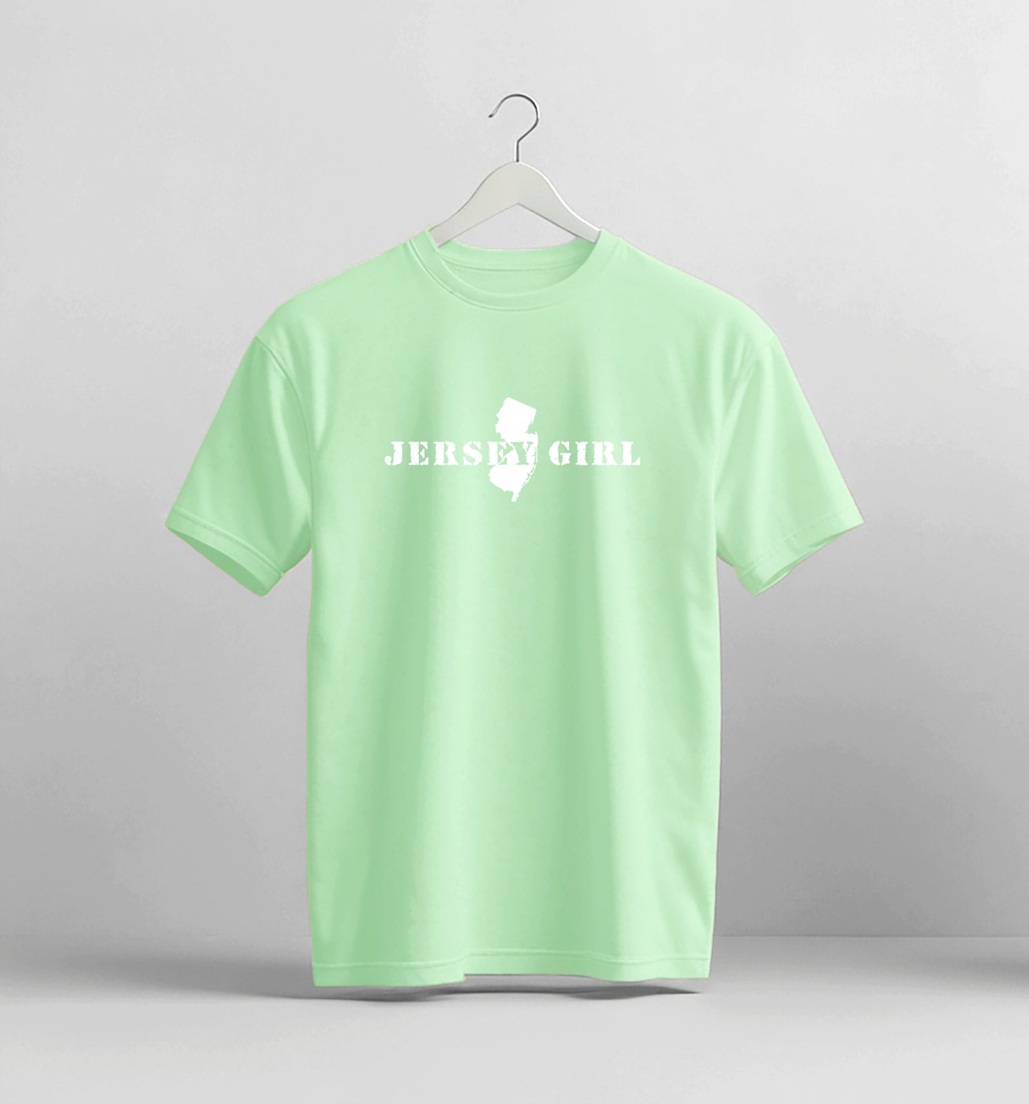 Light green t-shirt with 'Jersey Girl' text and map graphic on a hanger against a gray background